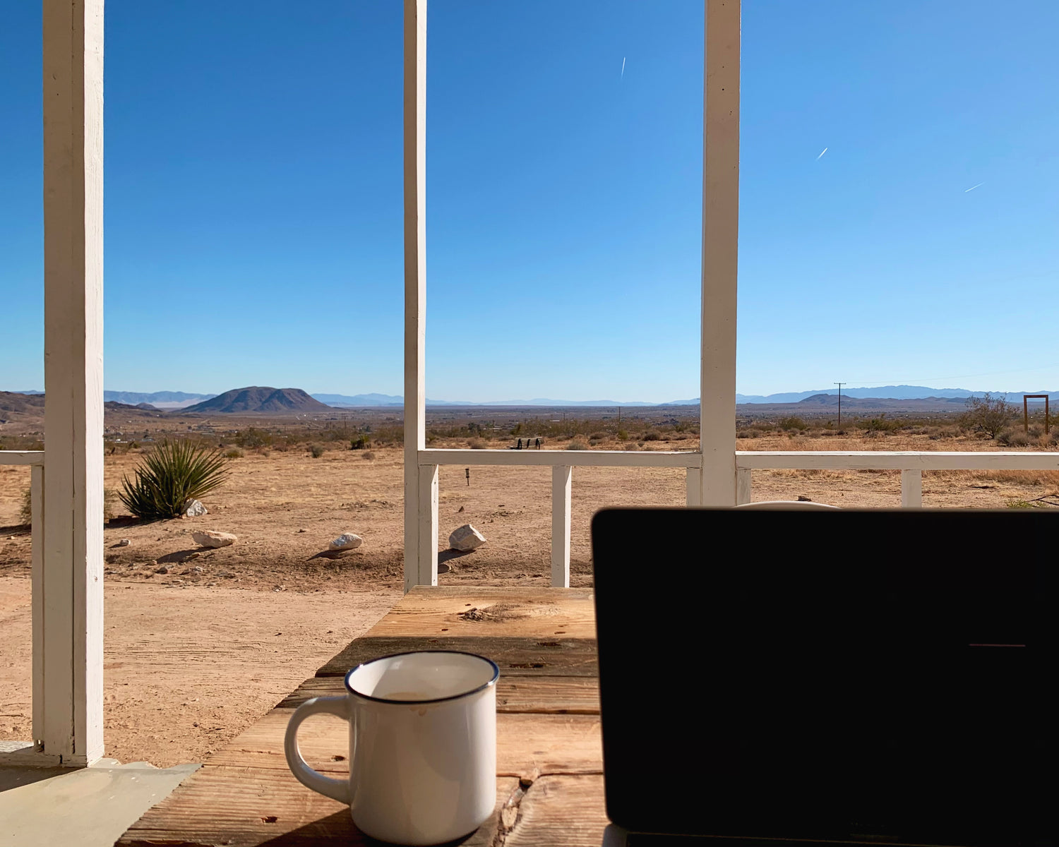 Laptop on a wooden desk with a view of a desert landscape through large windows.