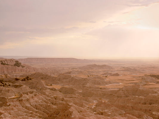 A serene Badlands landscape just before rainfall, with layered rock formations and a softly glowing sky that holds the stillness of an approaching storm.